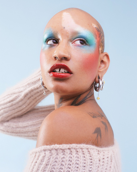 portrait of a bald person with tattoos on their head and back wearing watercolor coral and blue makeup and an...